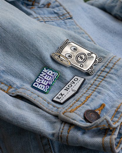 three custom enamel pins near the button cuff of a light denim jacket; top left pin features stacked text "Drink Beer Do Good" in white lettering with shiny rainbow finish around; right pin is large silver brew tank with details of manway hatch, zwickel spout, legs, coiled hose, etc. and large white googly eyes; bottom left pin is white chevron shape with black border and black inset lettering large "Ex Novo" and small "Brewing Company" beneath