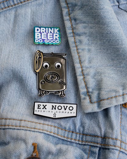 three custom enamel pins stacked vertically near the collar of a light denim jacket; top pin features stacked text "Drink Beer Do Good" in white lettering with shiny rainbow finish around; center pin is large silver brew tank with details of manway hatch, zwickel spout, legs, coiled hose, etc. and large white googly eyes; bottom pin is white chevron shape with black border and black inset lettering large "Ex Novo" and small "Brewing Company" beneath
