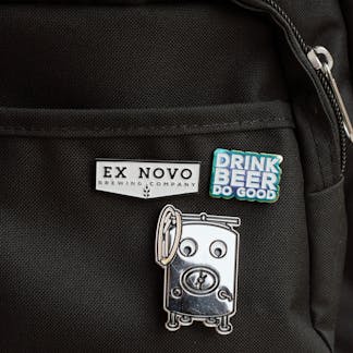 three custom enamel pins on a black canvas backpack near pocket and zipper; top right pin features stacked text "Drink Beer Do Good" in white lettering with shiny rainbow finish around; center bottom pin is large silver brew tank with details of manway hatch, zwickel spout, legs, coiled hose, etc. and large white googly eyes; top left pin is white chevron shape with black border and black inset lettering large "Ex Novo" and small "Brewing Company" beneath