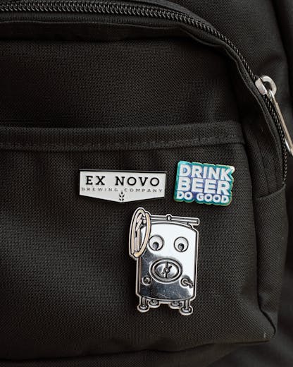 three custom enamel pins on a black canvas backpack near pocket and zipper; top right pin features stacked text "Drink Beer Do Good" in white lettering with shiny rainbow finish around; center bottom pin is large silver brew tank with details of manway hatch, zwickel spout, legs, coiled hose, etc. and large white googly eyes; top left pin is white chevron shape with black border and black inset lettering large "Ex Novo" and small "Brewing Company" beneath