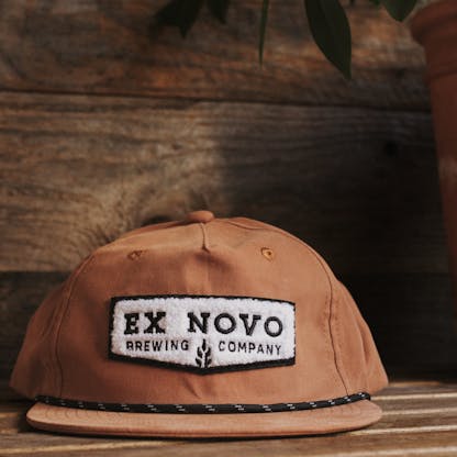 rust orange baseball hat with black rope along bill and fluffy embroidered chenille chevron shaped logo with black lettering "Ex Novo Brewing Company" against wood backdrop