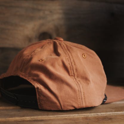 three-quarter back view of rust orange colored baseball hat with black plastic snapback closure against wood background