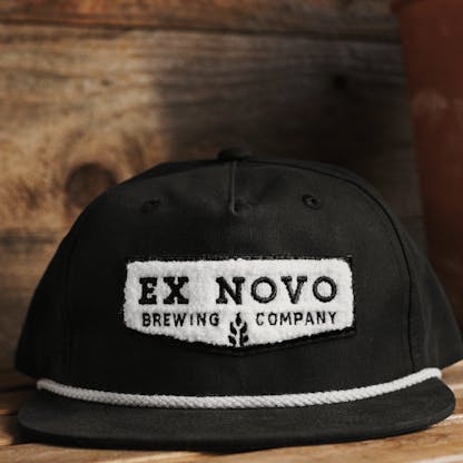 front view of black baseball hat with white rope along bill and white embroidered chenille chevron shaped logo with black lettering inside "Ex Novo Brewing Company" against wood background