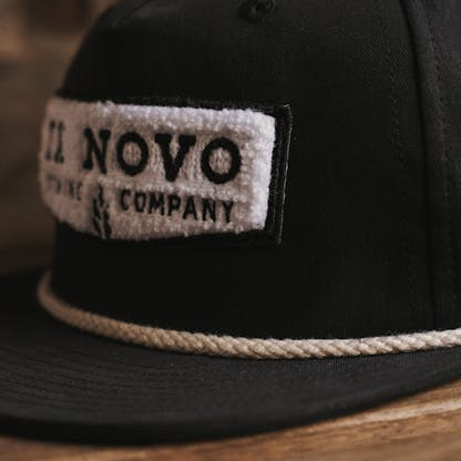 three-quarter front view of black baseball hat with white rope along bill and close up of texture on white emrboidered chenille chevron shaped logo with inset black lettering "Ex Novo Brewing Company"