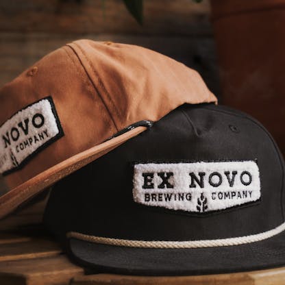 two baseball hats against wood background, rust colored orange with black rope along bill propped on top of black hat with white rope along bill. both hats feature white embroidered chenille chevron shaped logos on the front with black inset lettering "Ex Novo Brewing Company"