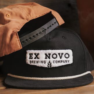 two baseball hats against wood background, backview of rust colored orange hat with black plastic snapback closure propped on top of front view of black hat with white rope along bill, featuring white embroidered chenille chevron shaped logos on the front with black inset lettering "Ex Novo Brewing Company"