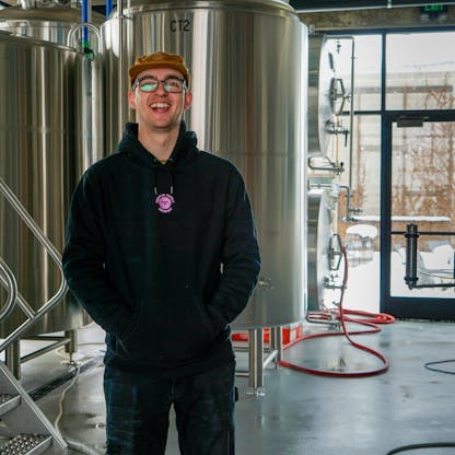 Male wearing black hoodie with lilac embroidered T.F. logo on upper center chest standing in a brewery.