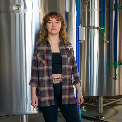 Female wearing plaid long sleeve flannel in brown and deep red tones with embroidered T.F. logo standing in front of a beer conditioning tank.