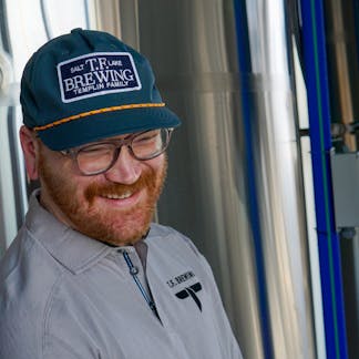 Male with a beautiful smile wearing a not quite blue but not quite gray hat with a NAVY TF Brewing logo patch with white writing on center.