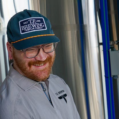Male with a beautiful smile wearing a not quite blue but not quite gray hat with a NAVY TF Brewing logo patch with white writing on center.