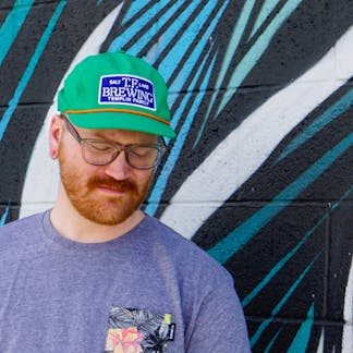 Male wearing a kelly green hat with a navy TF Brewing logo patch with white writing on the center standing in front of a mural.