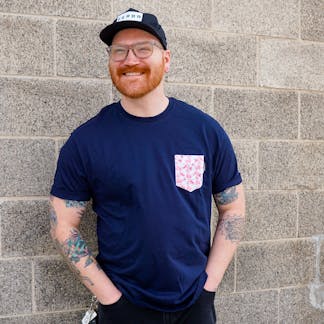 Male wearing a navy t-shirt with a T.F. beer label graphic pocket design. The pocket has pink lingonberry flowers. Pairs well with our lingonberry berliner weisse.