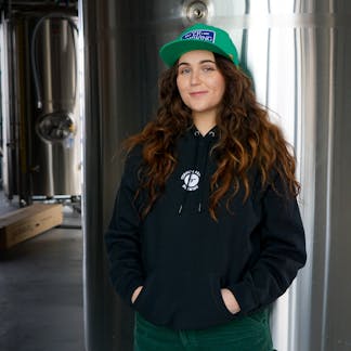 Female wearing black hoodie with white embroidered T.F. logo on upper center chest standing in a brewery.