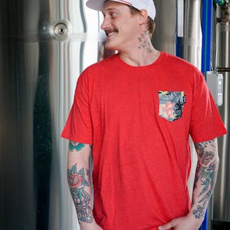 male wearing red t-shirt with beer label graphic pocket on upper left chest. The design mimics our coconut guava Berliner weisse can art. pink Hibiscus flower, gray scale plant life.