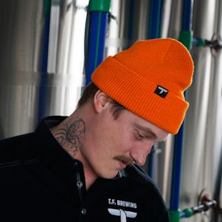 Male wearing a neon orange beanie with a T.F. logo tag standing in a brewery.
