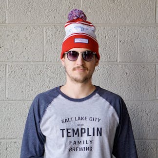 Male wearing a red, white and blue T.F. logo pom pom beanie standing in front of a wall.
