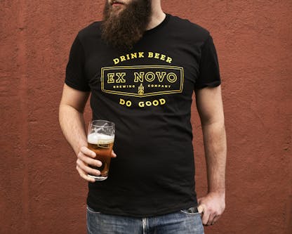 close up view of a bearded man's torso standing in front of a red wall holding a partial pint of beer and looking off in the distance. he wears a black t-shirt with yellow text "Drink Beer Do Good" arching above and below a chevron logo with internal text "Ex Novo Brewing Company"