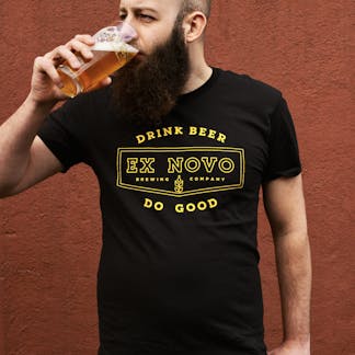 a bearded man stands in front of a red wall drinking a beer and looking off in the distance. he wears a black t-shirt with yellow text "Drink Beer Do Good" arching above and below a chevron logo with internal text "Ex Novo Brewing Company"