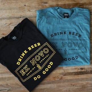 two t-shirts folded square on a wood surface; black with yellow chevron design and text "Drink Beer Do Good" arching above and below "Ex Novo Brewing Company"; tucked beneath and to the right is a teal t-shirt with dark green text in the same design