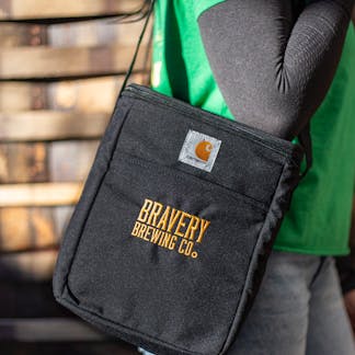 Bravery Carhartt beer cooler. The cooler is black fabric on the outside with the text "Bravery Brewing Co." embroidered in yellow. The model is wearing the cooler by its long strap, hanging from their shoulder.