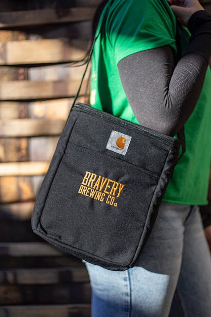 Bravery Carhartt beer cooler. The cooler is black fabric on the outside with the text "Bravery Brewing Co." embroidered in yellow. The model is wearing the cooler by its long strap, hanging from their shoulder.