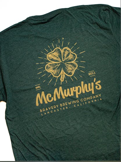 back of a dark green heather McMurphy's t-shirt. McMurphy's logo art is printed large on the back in yellow ink along with the text " McMurphy Bravery Brewing Company Lancaster, California. Est. 2011"