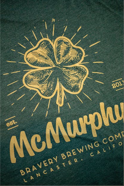 detail of the back of a dark green heather McMurphy's t-shirt. McMurphy's logo art is printed large on the back in yellow ink along with the text " McMurphy Bravery Brewing Company Lancaster, California. Est. 2011"