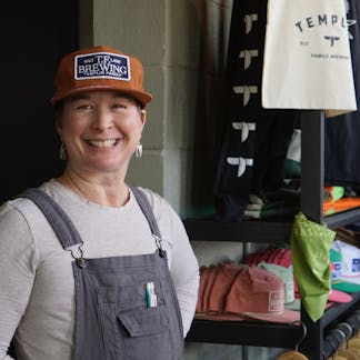 Female in overalls with a bright smile wearing a rusty clay brown hat with a navy TF Brewing logo patch with white writing on the center.