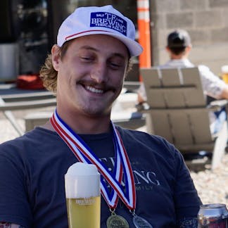 Male decorated in beer competition medals wearing a white hat with a navy TF Brewing logo patch with white writing on the center of the hat.