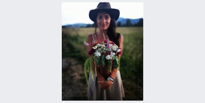 Woman standing in a field holding a bouquet of flowers. She will be offering a 90 minute flower workshop and one beer is included in the price.