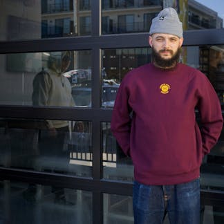 Male wearing a Maroon crewneck with gold embroidered T.F. logo on upper center chest standing in front of glass panel garage door.