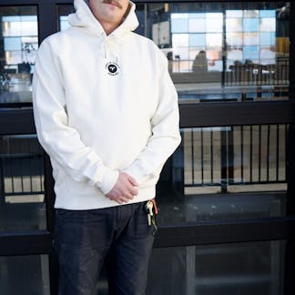 Male wearing cream hoodie with embroidered T.F. logo on upper center chest standing in front of glass panel garage door with reflection of the street.