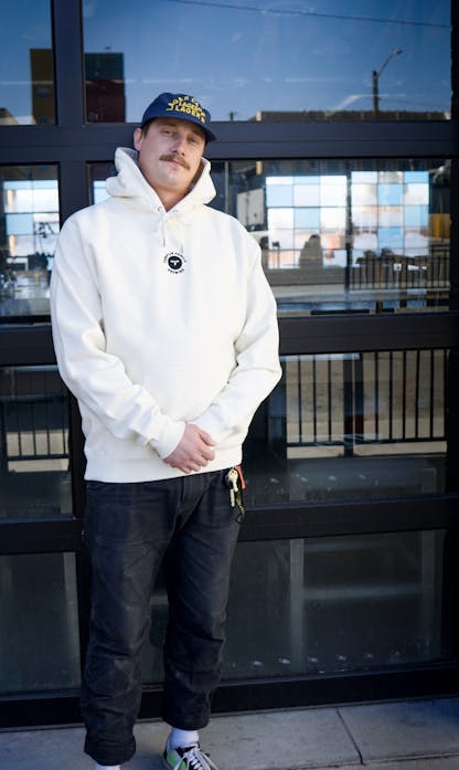 Male wearing cream hoodie with embroidered T.F. logo on upper center chest standing in front of glass panel garage door with reflection of the street.