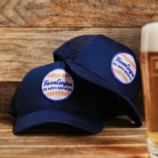 two navy hats, one atop the other, with mesh back and front white, red, and blue baseball patch with cursive text "Farm League" and block text "Ex Novo Brew Co"; to the right a pint of beer can be glimpsed