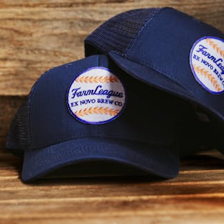 two navy hats, one atop the other, with mesh back and front white, red, and blue baseball patch with cursive text "Farm League" and block text "Ex Novo Brew Co"