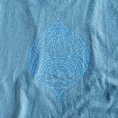 Close up of Milagro graphic with clouds, rainbows, and sun rays in light blue on a denim wash material
