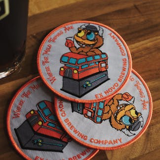 Three circular gray patches with a red border piled together of Where the Mild Things are character riding in a red London bus holding a pint glass