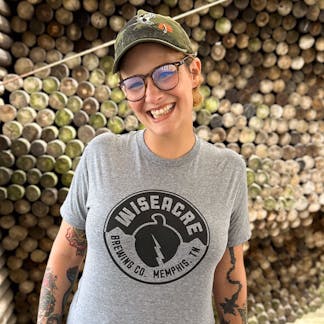 A person smiling modeling a grey shirt with the Wiseacre Brewing Company Acorn Logo in black across the chest