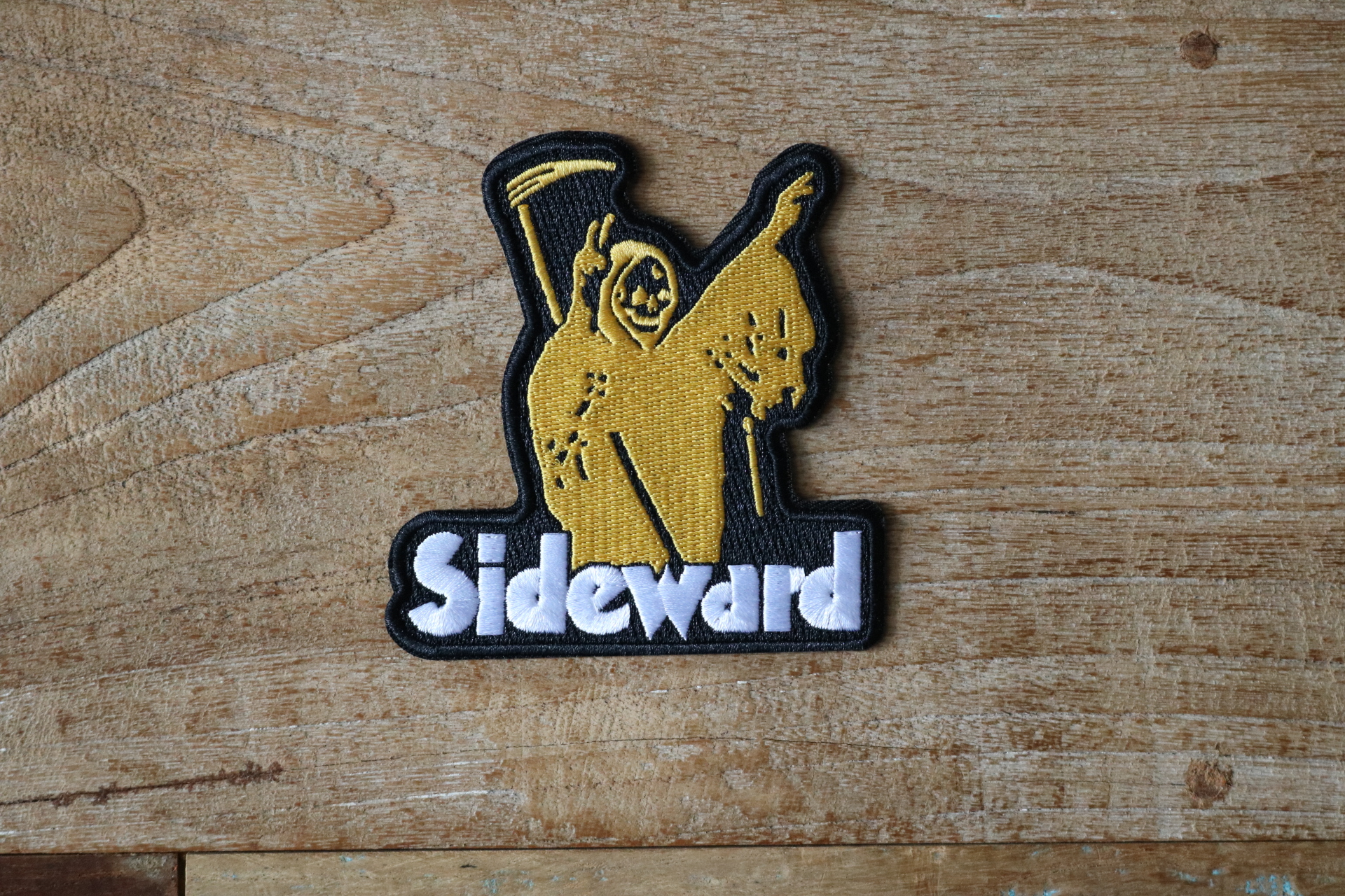 An embroidered black patch featuring a yellow grim reaper holding peace signs with the text "Sideward" underneath.