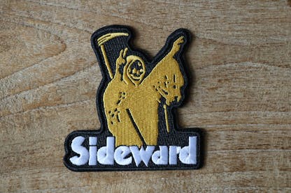 Detailed image of an embroidered black patch featuring a yellow grim reaper holding peace signs with the text "Sideward" underneath.