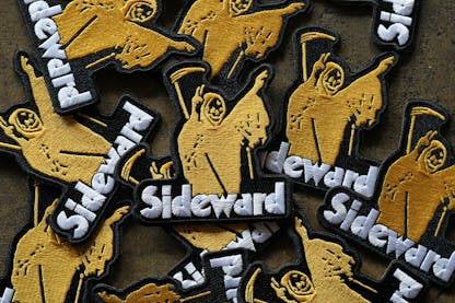 A pile of embroidered black patches featuring a yellow grim reaper holding peace signs with the text "Sideward" underneath.