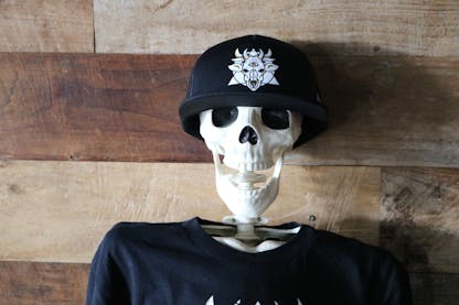 A plastic skeleton wearing a black "trucker-style" hat. The front of the hat has the Sideward logo which resembles a many horned demon cow skull with a triangle and circle behind it.