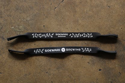 One neoprene sunglasses holder features the Sideward Brewing wordmark surrounded by a lot of beer cans. The second neoprene sunglasses holder features fewer beer cans and larger Sideward Brewing wordmark, with their secondary logo, an eye, in the center.