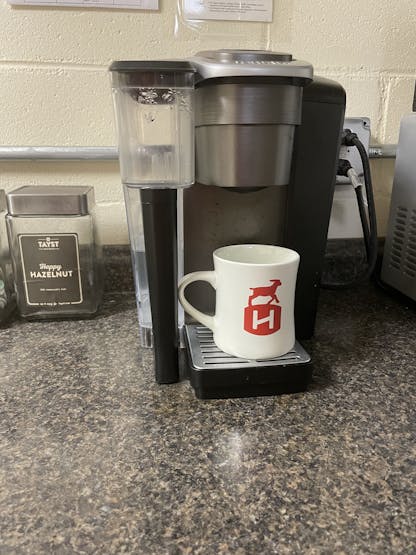 cream mug with red hardywood sheep and barrel logo on the side sits under a keurig coffee maker