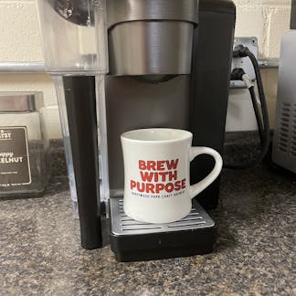 cream coffee mug with brew with purpose text in red on the side sitting under a keurig coffee maker