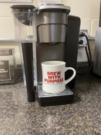 cream coffee mug with brew with purpose text in red on the side sitting under a keurig coffee maker