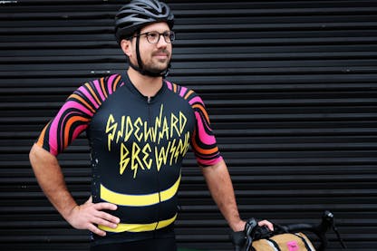 Austin models our zippered cycling jersey - the front design is handdrawn letters reading Sideward Brewing in yellow on a black jersey with pink and orange wavy striped sleeves.