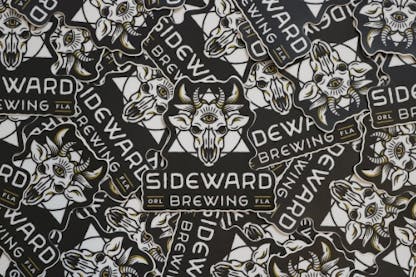A pile of stickers of Sideward Brewing's logo, which resembles a demon cow skull with a triangle and circle behind it, with the Sideward Brewing wordmark below.