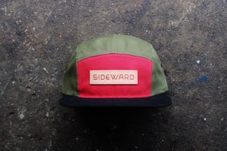 A five paneled hat, where four of the panels are olive green, the front facing fifth panel is red with a leather patch reading Sideward, with a black bill.