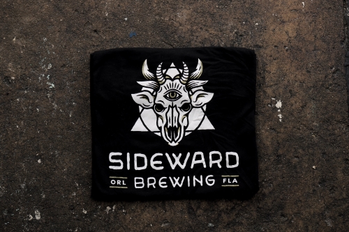 A black tee featuring a white Sideward Brewing logo, which resembles a demon cow skull with a triangle and circle behind it, with the Sideward Brewing wordmark below.